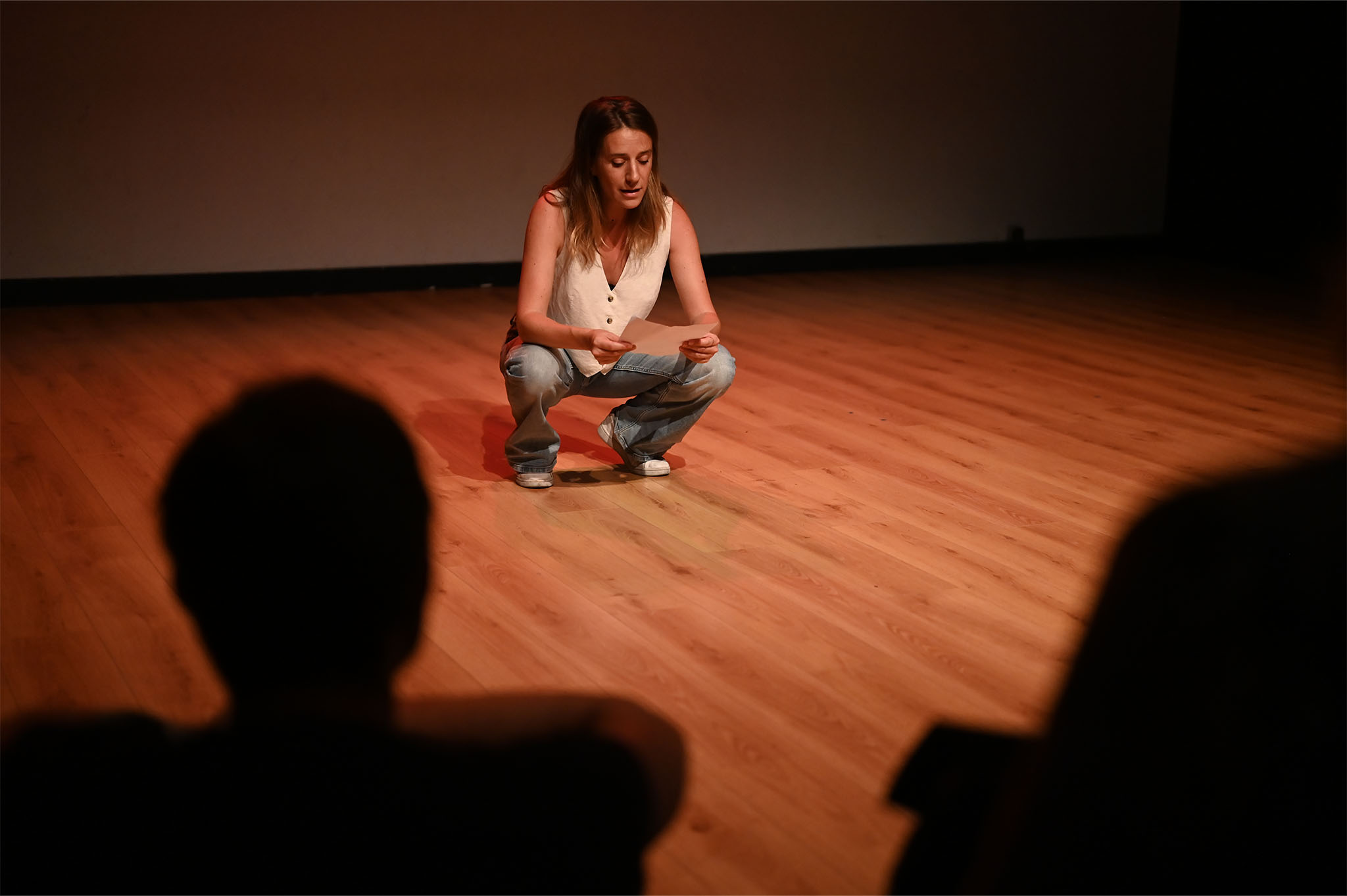 Au premier plan, des gens de dos regardent une femme accroupie en train de lire un texte sur le plateau d'un théâtre